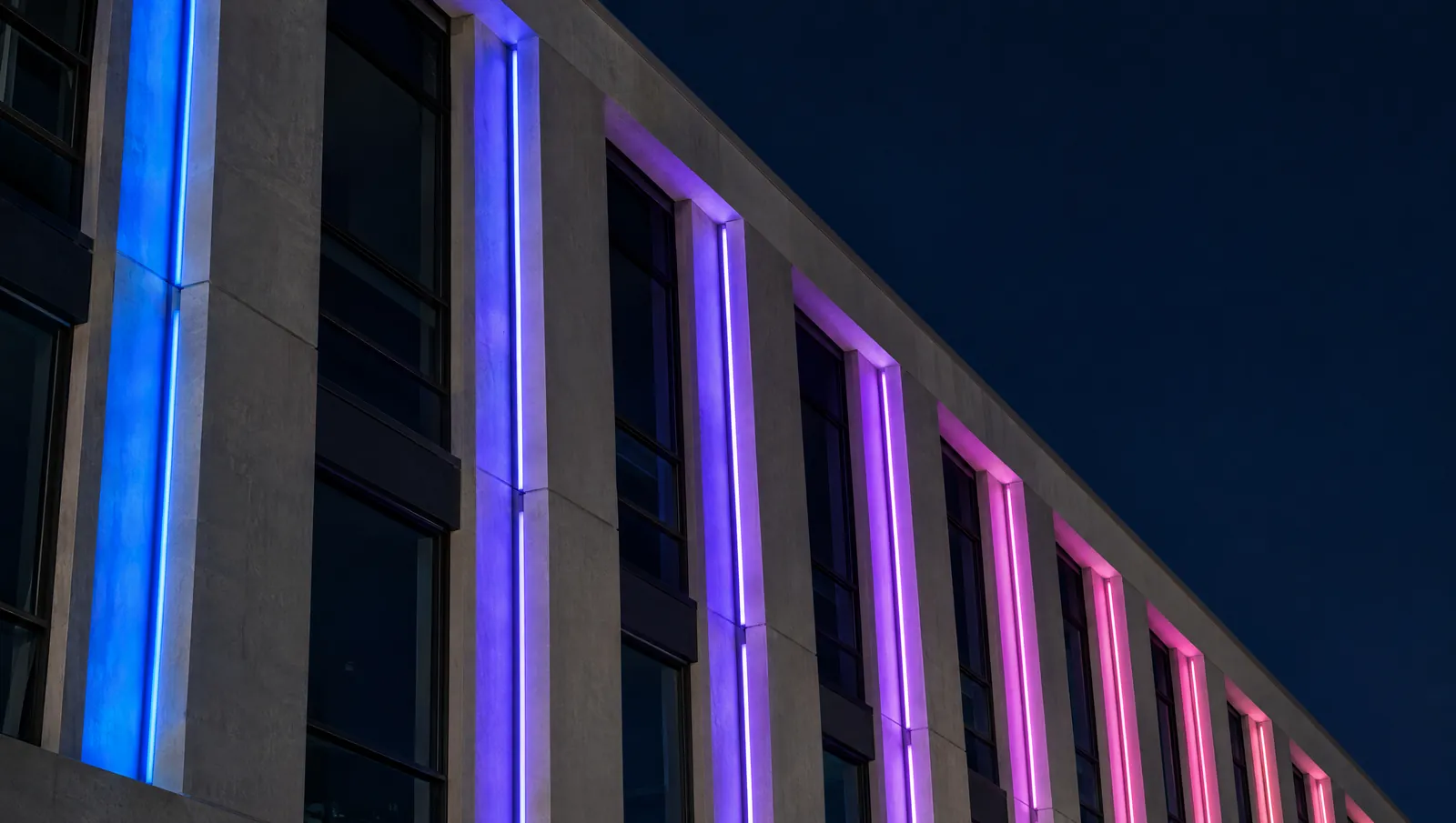 Facade at night with a linear luminaire in each window bay, each bay a different colour in one static sequence from blue to magenta